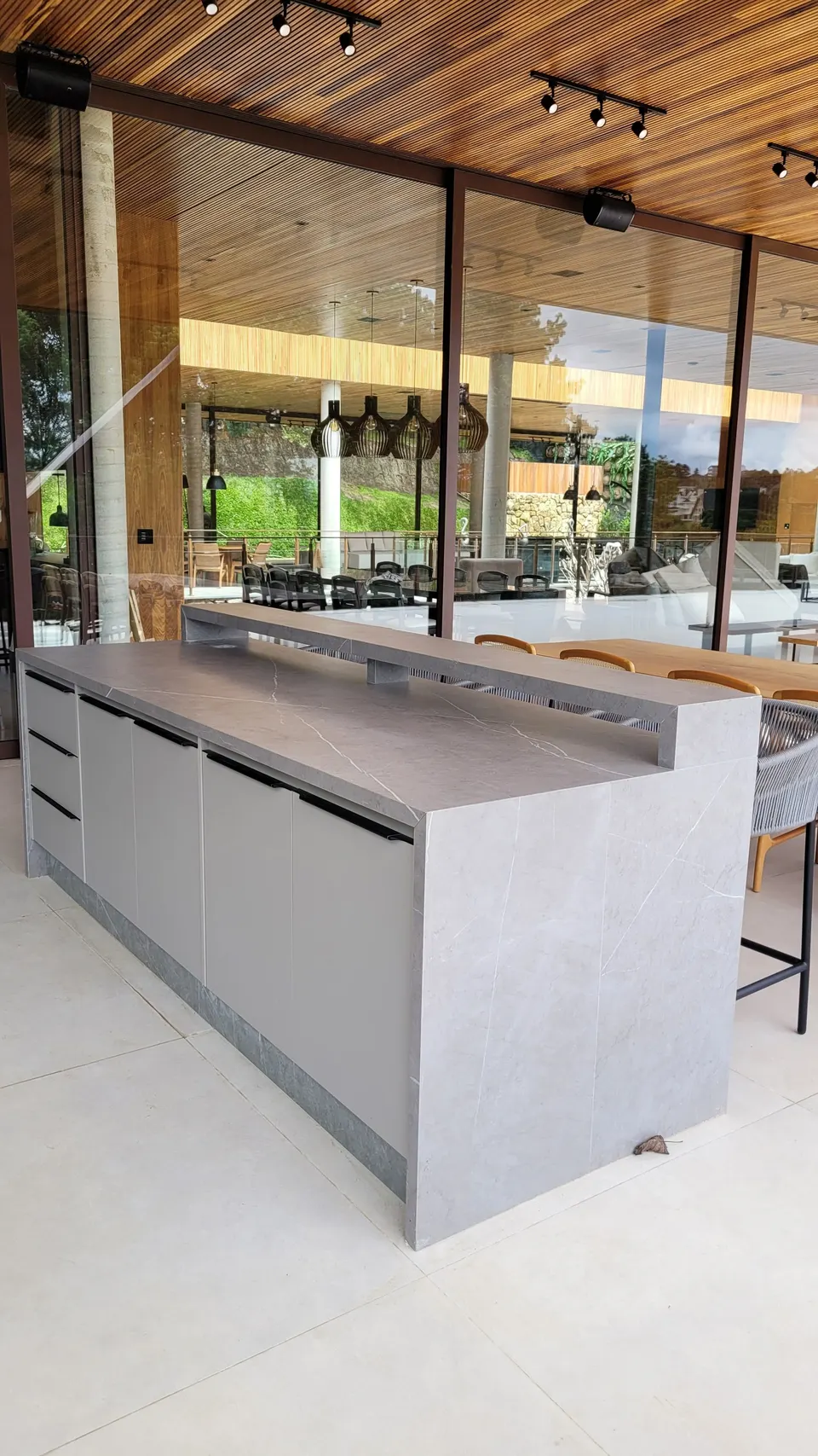 Ilha de cozinha em lâmina cinza claro em área gourmet integrada com amplas janelas e vista para o exterior