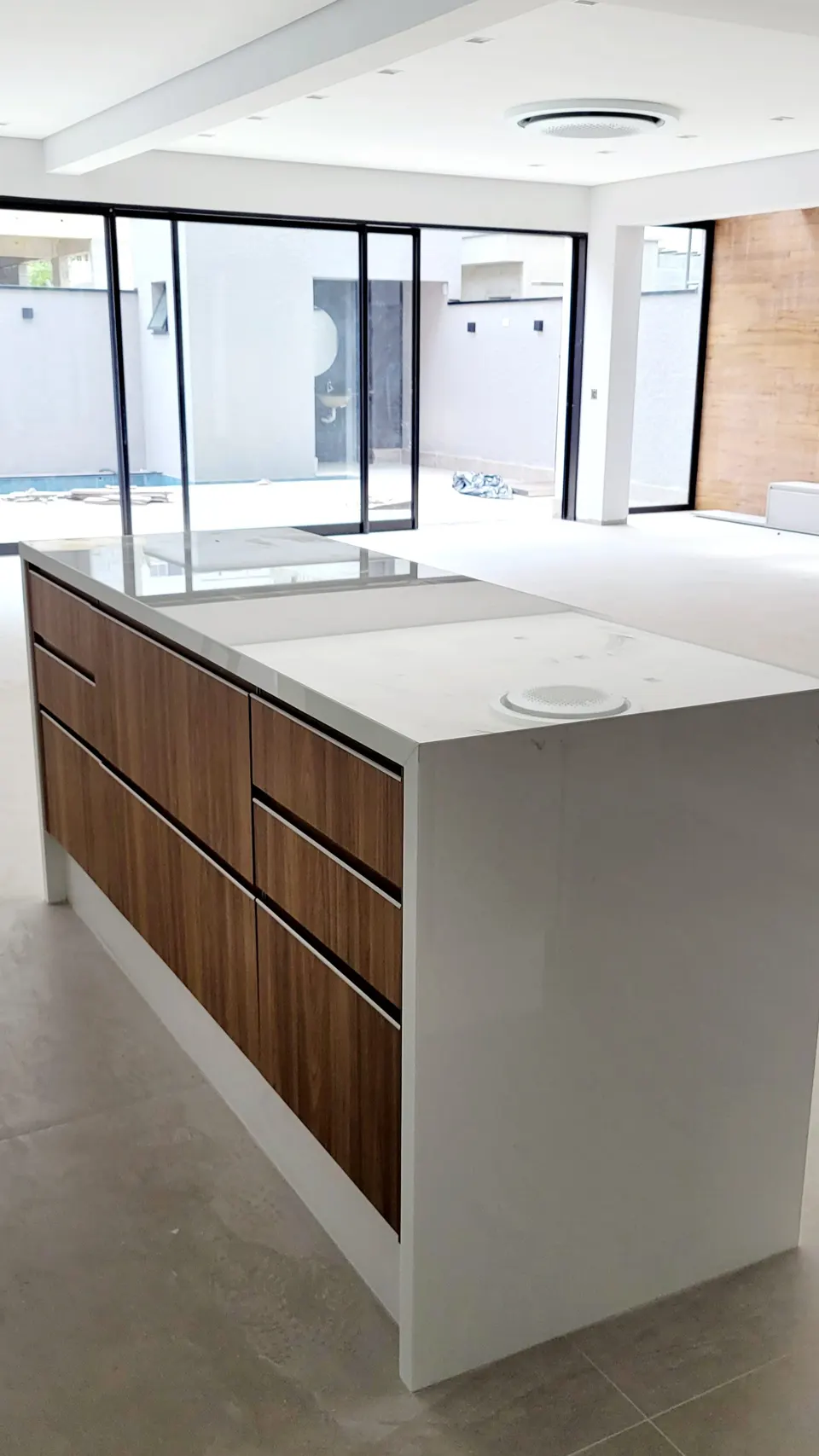 Ilha de cozinha em quartzo branco com acabamento polido, estrutura de madeira escura, dois cubas/espaços integrados na superfície, em ambiente moderno com pé-direito alto e amplas aberturas de vidro
