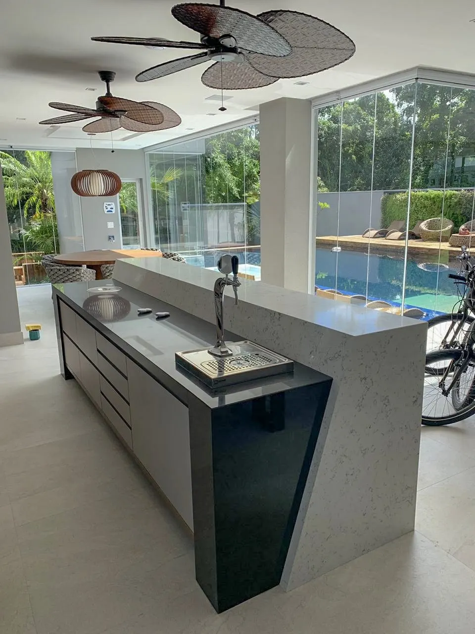 Bancada de área gourmet em pedra clara com acabamento polido, integrada à estrutura de bar junto a grandes janelas com vista para piscina — Granito Preto Absoluto