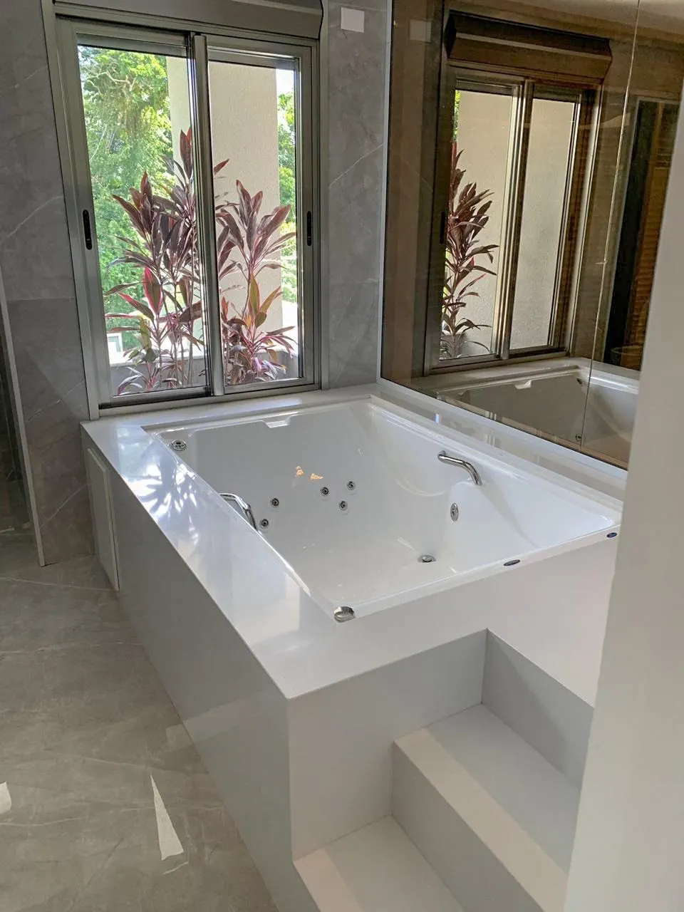 Banheira branca com acabamento em pedra branca, estrutura em alvenaria com revestimento de piso cinza claro no banheiro — Prime Branco