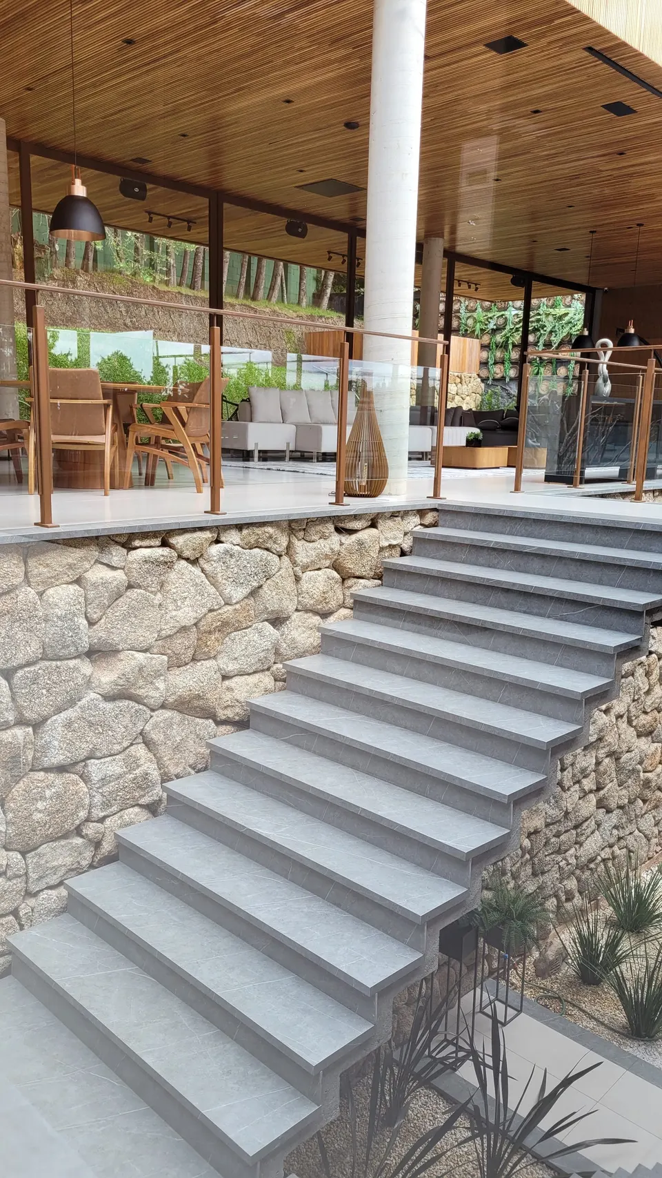 Escada externa em lâmina cinza com degraus, integrada a revestimento de parede em pedra natural clara e piso em tom neutro